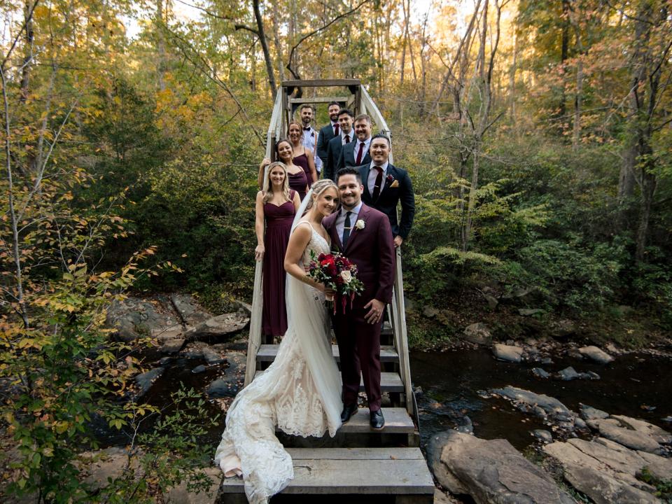 Bride, groom, and wedding party posing for a photo on a bridge over a creek