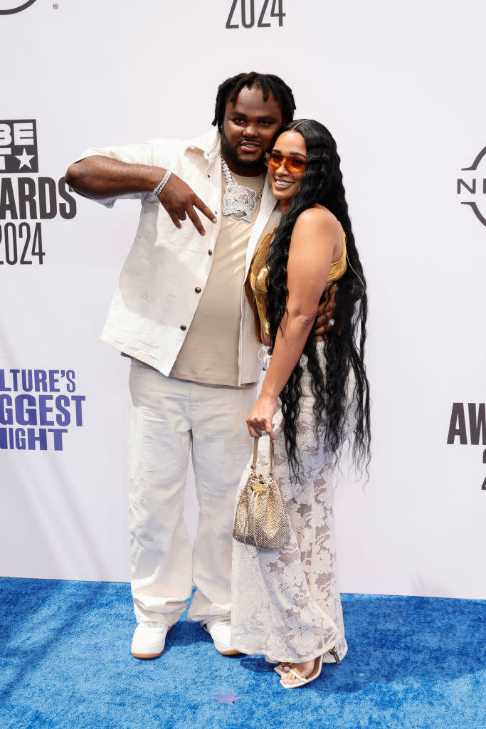 Celebrities Tee Grizzley and his wife, My'Eisha Agnew, pose on a red carpet. Tee wears a casual white outfit, while My'Eisha wears a stylish lace dress and sunglasses