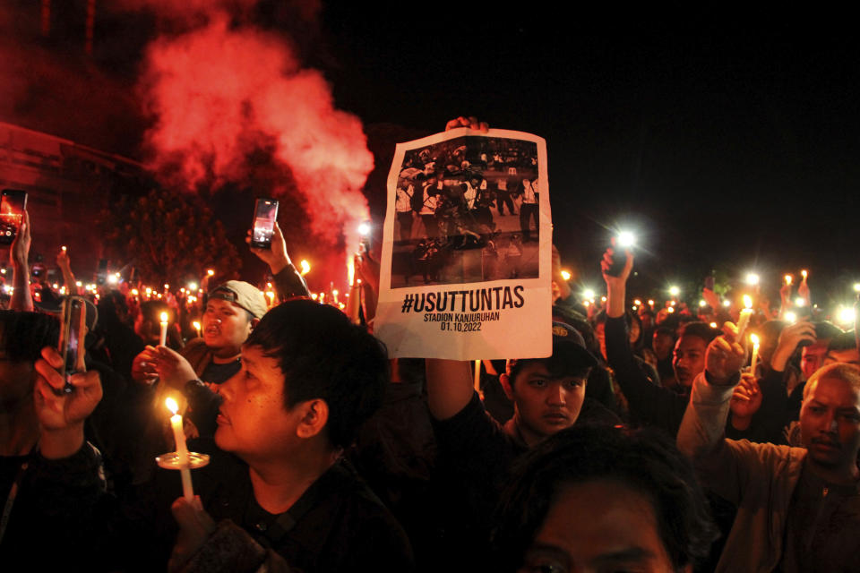 FILE - Soccer fans light a flare during a candle light vigil for the victims of Saturday's stampede, in Yogyakarta, Indonesia, Tuesday, Oct. 4, 2022. Police firing tear gas inside a stadium in East Java on Saturday in an attempt to stop violence after an Indonesian soccer match triggered a disastrous crush of fans making a panicked, chaotic run for the exits, leaving at a number of people dead, most of them trampled upon or suffocated. Writing on the poster reads "Investigate thoroughly". (AP Photo/Slamet Riyadi, File)