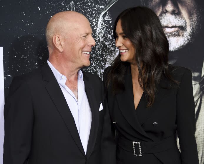 A bald man and a woman with long, brown hair smiling at each other while wearing black suits