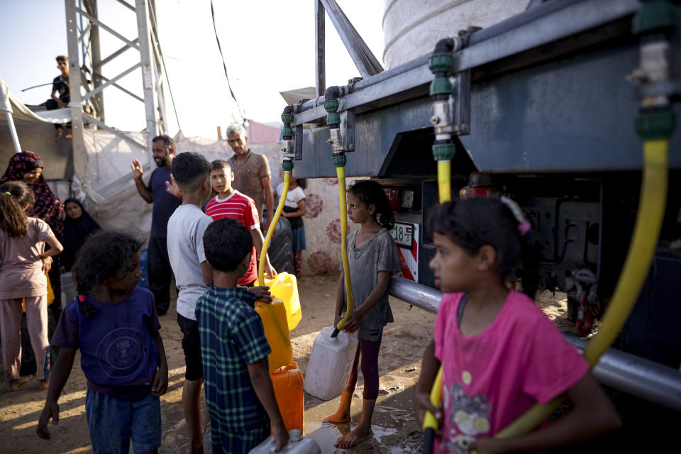 Palestinian children displaced by the Israeli bombardment of the Gaza Strip fill plastic bottles with water as others queue and wait their turn at a makeshift tent camp in the southern town of Khan Younis, Monday, July 1, 2024. (AP Photo/Jehad Alshrafi)