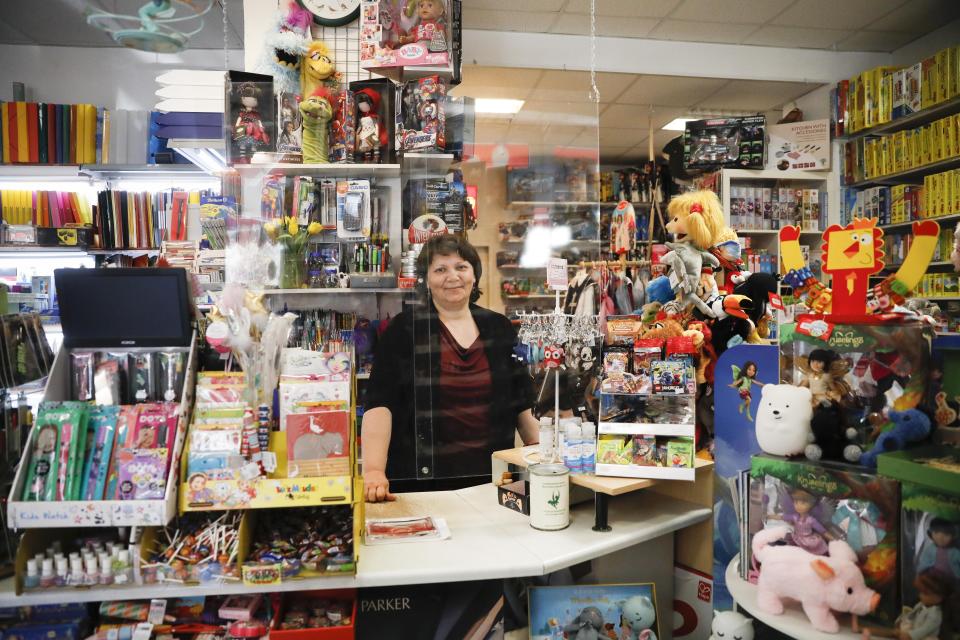 Galina Hooge owner of a small stationery and toy store poses for a photo behind plexiglass, to protect against the coronavirus, in her shop in Berlin, Germany, Wednesday, April 22, 2020. Galina Hooge opened her store for the first time in over a month, welcomed the loosening of restrictions, but remained wary. (AP Photo/Markus Schreiber)