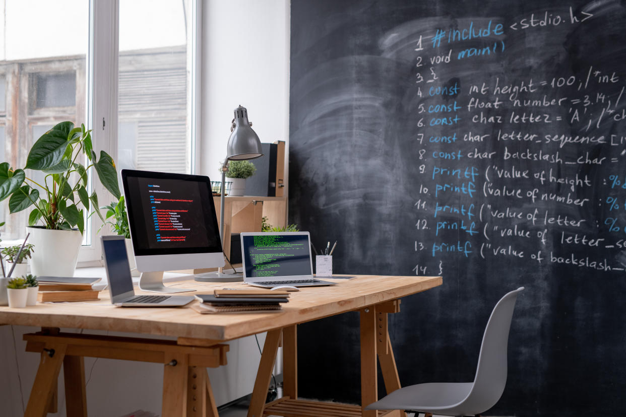 Workplace of it engineer by office window with two laptops, computer monitor, some books and copybooks, lamp and domestic plants