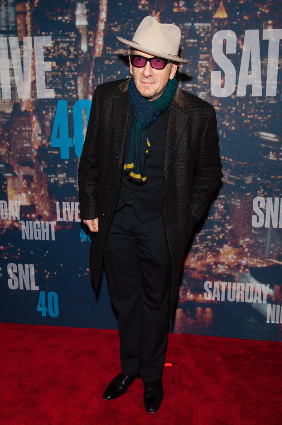 Elvis Costello must secretly be attending New York Fashion Week — or following along at home — because his outfit, from the wide-brimmed hat, to plaid coat, and tinted glasses, are just so trendy.