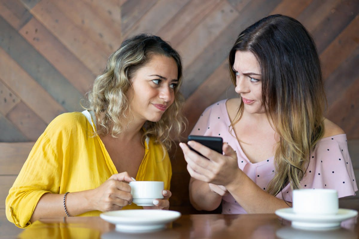 Two women look displeased as they have coffee and look at a phone. 