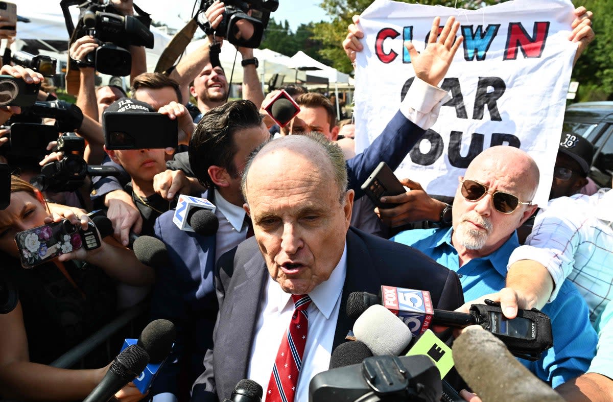 A person holds a sign reading ‘Clown Car Coup’ as  Rudy Giuliani speaks to the media after being booked at the Fulton County Jail in Atlanta, Georgia (AFP via Getty Images)