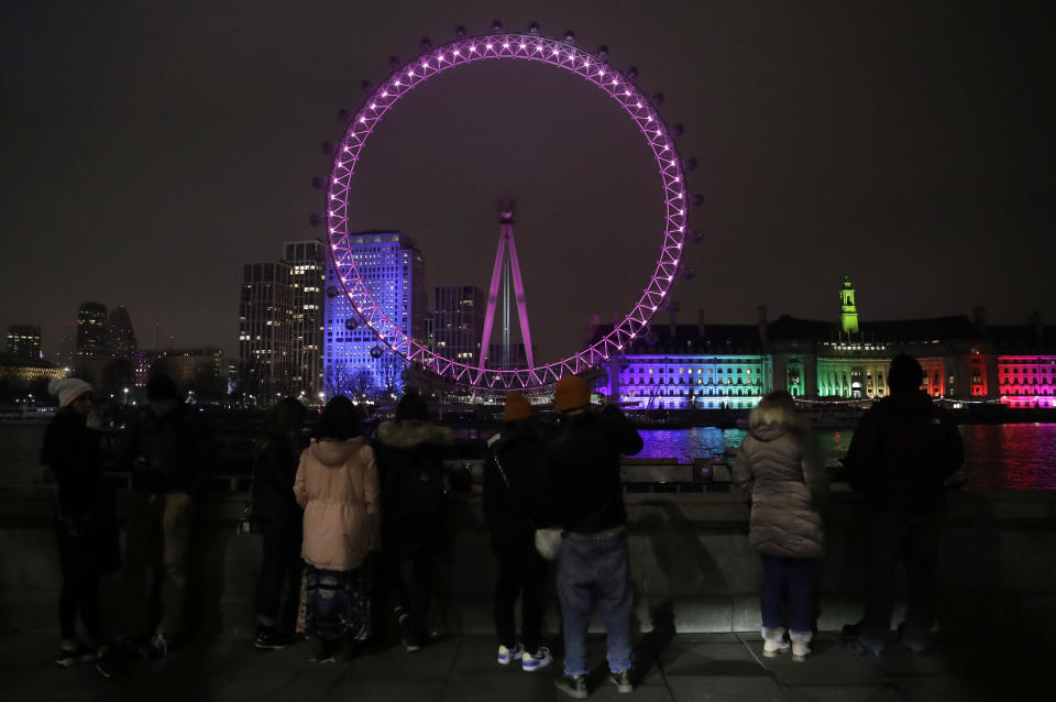 A small group of people look across from the embankment towards the London Eye ferris wheel by the River Thames in London, Thursday, Dec. 31, 2020. The London Eye is one of the traditional sites for New Year's Eve firework display, but it has been cancelled due to the ongoing coronavirus pandemic and the restrictions in place to try and stop its spread. (AP Photo/Matt Dunham)