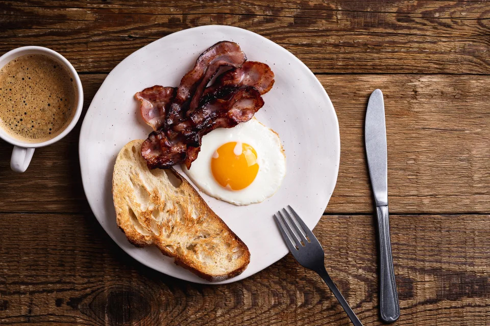 A plate with toast, a fried egg, strips of crispy bacon, and a cup of coffee on a wooden table