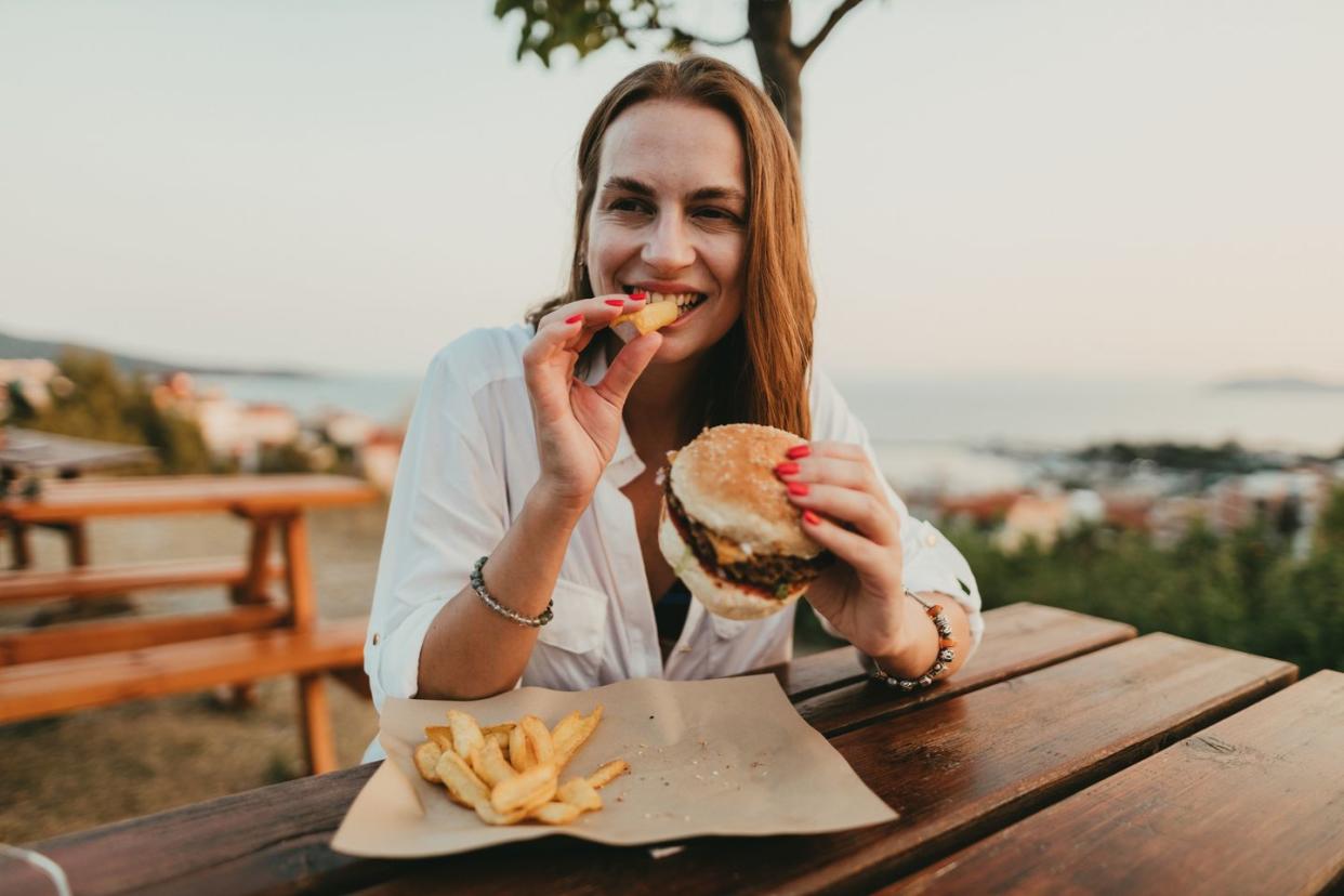 happy woman sitting outdoors and eating burger and fries on a sunny summer day during a seaside vacation