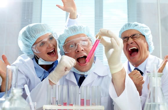Three scientists, mouths agape, in a laboratory looking at a test tube filled with pink liquid and celebrating.