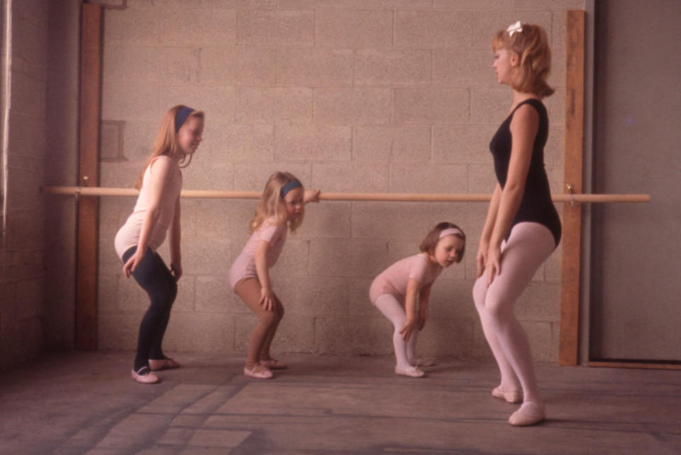 A ballet instructor in fitness wear teaches three young girls, each in ballet attire, how to plie in front of a barre