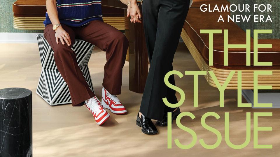 basketball star sue bird in burgundy pants and blue striped top and curly hair and red sneakers looking up at soccer captain megan rapinoe in black pants and a white lacey top in their living room