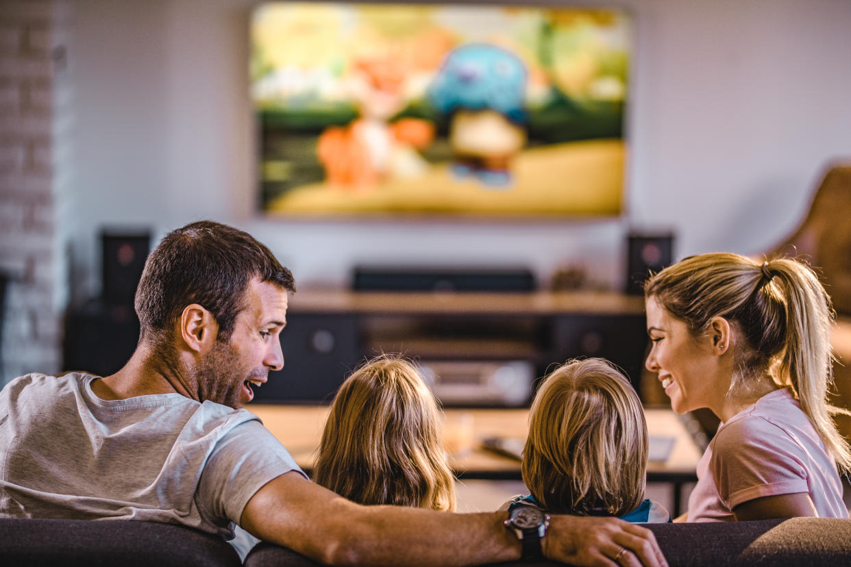 Back view of happy parents talking to their kids while watching TV on sofa in the living room.