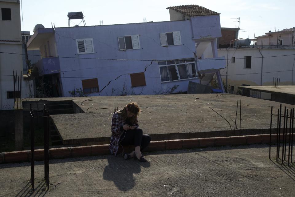 A woman checks her cellphone as a damaged building is seen in the background in Durres, western Albania, Wednesday, Nov. 27, 2019. The death toll from a powerful earthquake in Albania has risen to 25 overnight as local and international rescue crews continue to search collapsed buildings for survivors. (AP Photo/Visar Kryeziu)