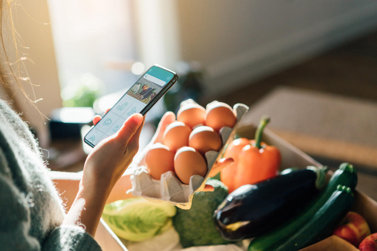 Cropped shot of woman using smartphone while picking up eggs from a delivery box filled with fresh organic fruits and vegetables at home.