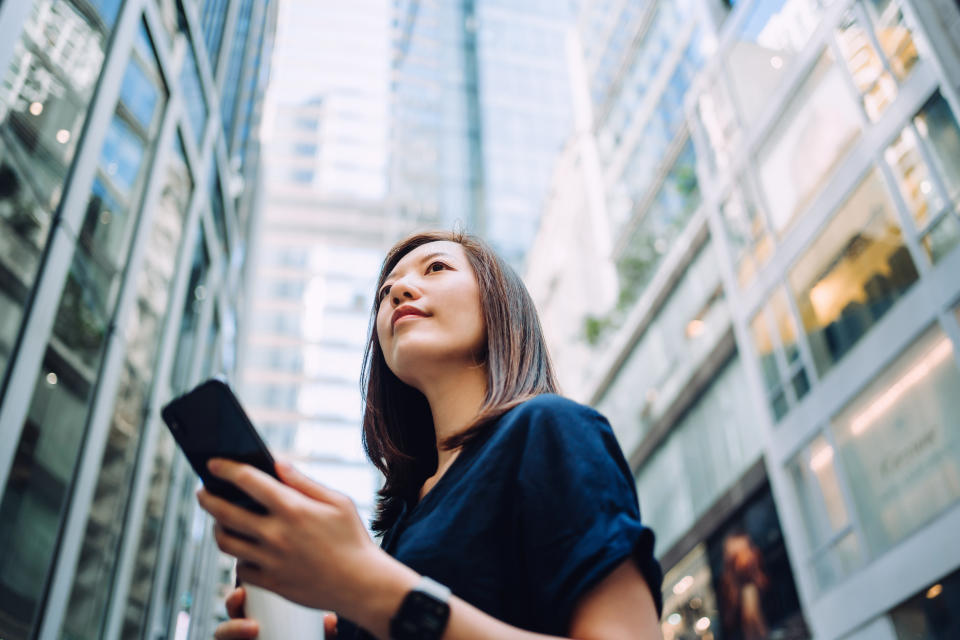 Young Asian businesswoman with coffee to go, using smartphone while standing against high-rise corporate buildings 