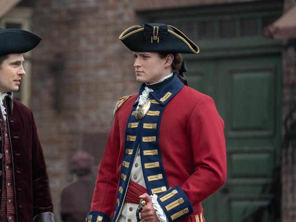 William Ransom wearing a three-peaked hat with golden trim and an ornate-looking red overcoat while staring at a man to his left.