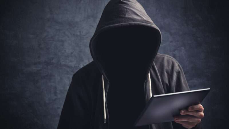 An anonymous man in a hoodie browses the internet on a tablet.