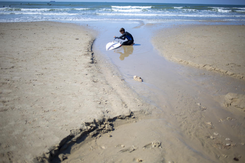 A surfer cleans his tar covered surfboard from an oil spill in the Mediterranean Sea in Gador nature reserve near Hadera, Israel, Saturday, Feb. 20, 2021. Hundreds of volunteers are taking part in a cleanup operation of Israeli shoreline as investigations are underway to determine the cause of an oil spill that threatens the beach and wildlife, at Gador Nature Reserve near the northern city of Hadera, the tar smeared fish, turtles, and other sea creatures. (AP Photo/Ariel Schalit)