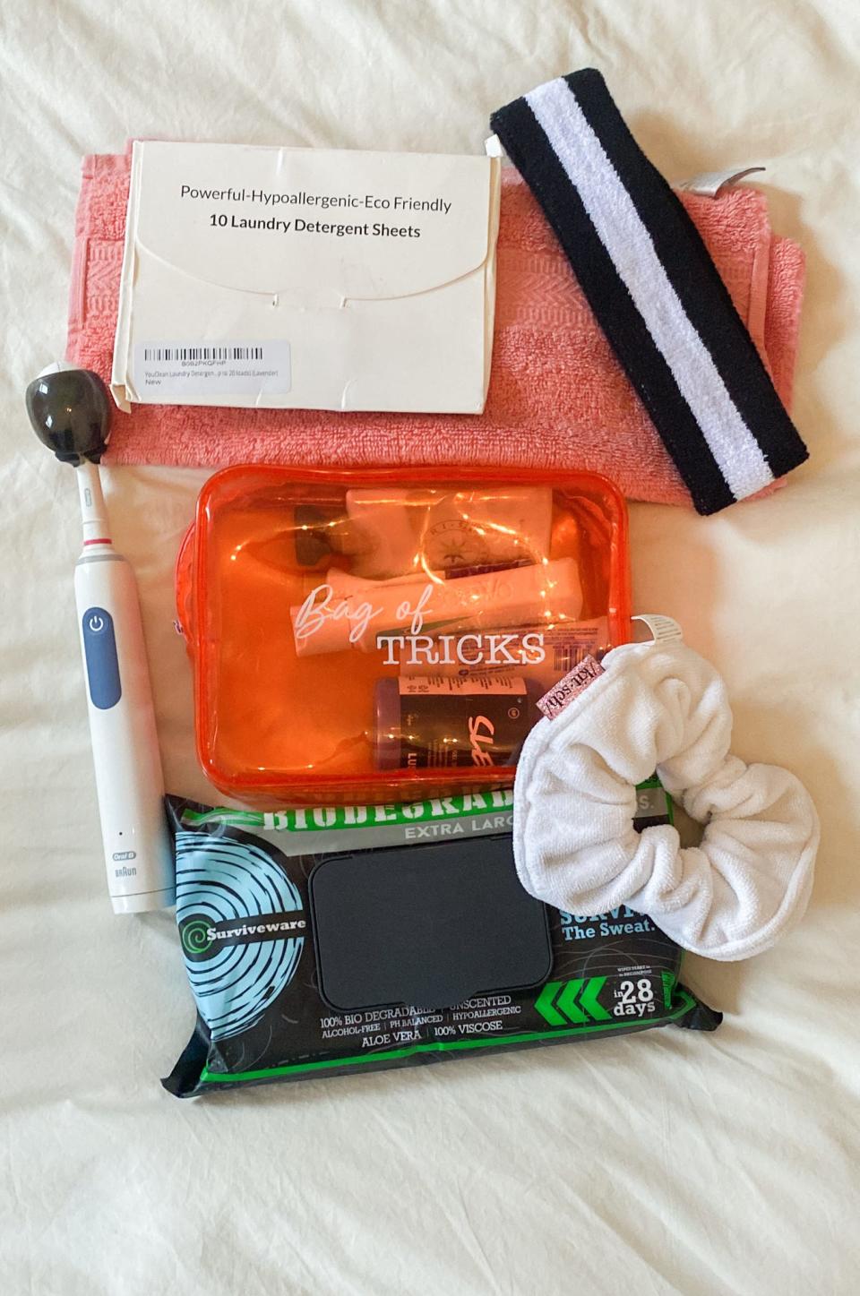 The author's toiletries on a white sheet including a sweatband and white package of laundry sheets on top of a pink rag. Below these items, there's a white and blue electric toothbrush with an astronaut helmet shaped cover, an orange bag of liquid items, a black package of wipes, and a thick, white scrunchy meant for drying hair.