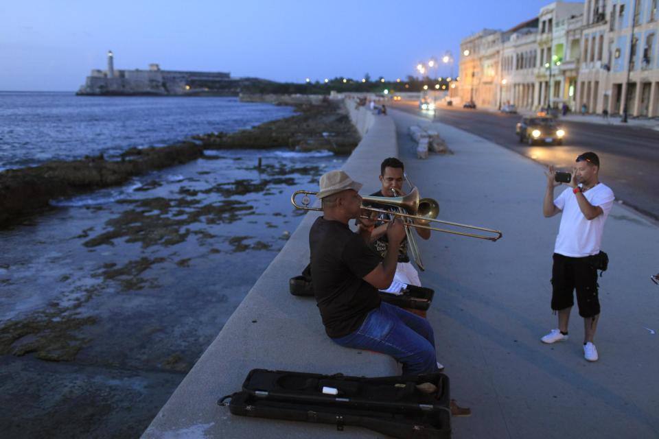 In this July 8, 2013 photo, musicians play trombones as a tourist from Colombia takes their picture along the Malecon in Havana, Cuba. For a place where most people earn just $20 a month at their government jobs, Havana can be a surprisingly expensive place to be a traveler. But there are plenty of free ways to have fun in this city known for sea, sun and salsa. (AP Photo/Franklin Reyes)