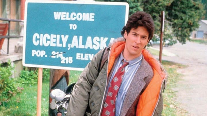 Joel standing in front of a sign that says welcome to Cicely, Alaska in a scene from Northern Exposure.