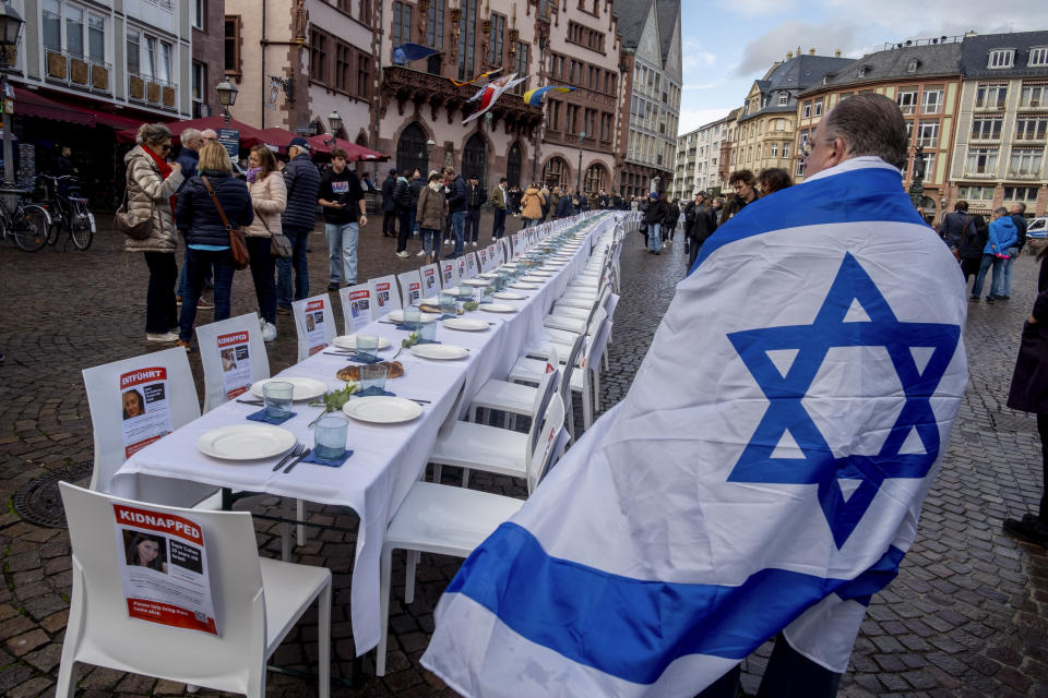 A man draped in an Israeli flag walks past a shabbat table with some 200 empty chairs in front of the town hall in Frankfurt, Germany, Friday, Oct. 27, 2023. The 200 empty chairs represent the Israeli hostages taken by Hamas. (AP Photo/Michael Probst)