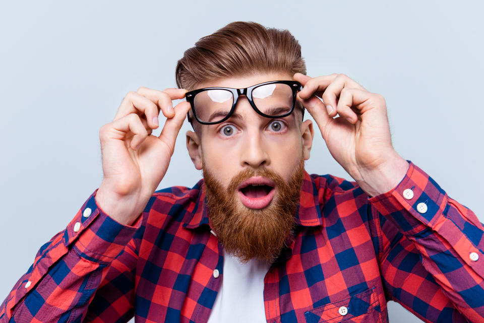 It's incredible! Close up portrait of young bearded man touching the spectacles and keeping his mouth open against gray background