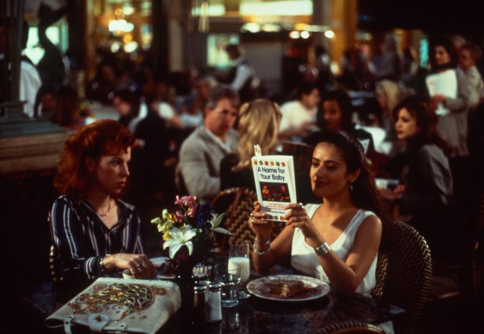 Hogan and Salma Hayek seated at a table in a restaurant. Hayek holds a book called: A Name for Your Baby.