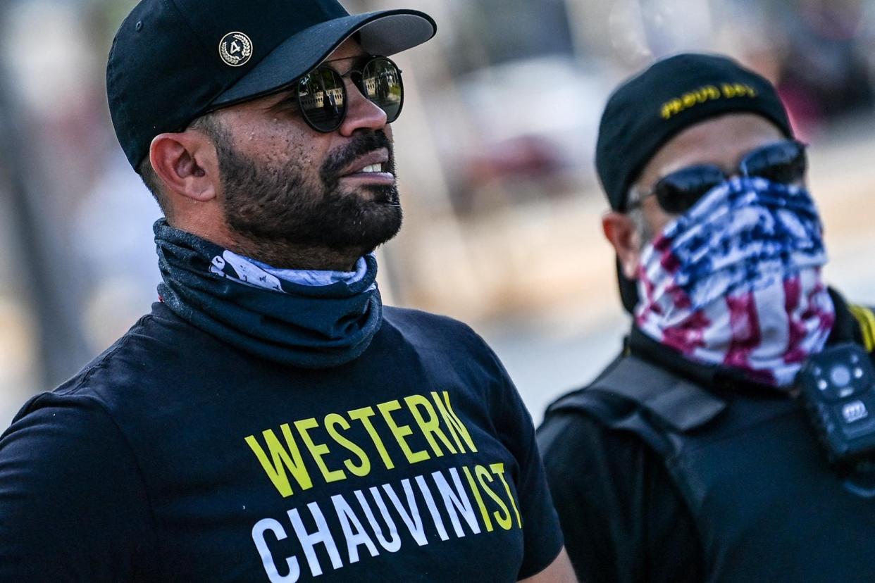 A bearded man wearing a black baseball hat, aviator sunglasses, a bandanna, and a black shirt that reads, "Western Chauvinist."