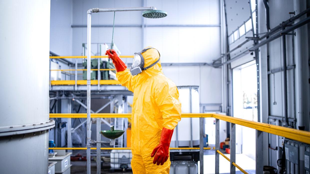  Person wearing a yellow biohazard suit in a decontamination shower against an industrial background. 