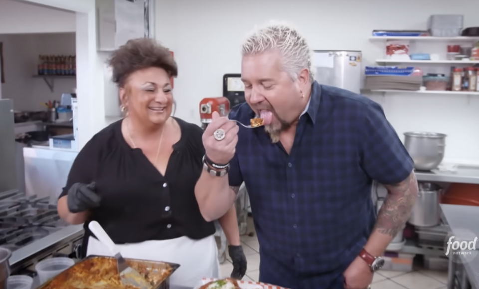 Woman feeds a man in a plaid shirt a sample of food in a kitchen setting