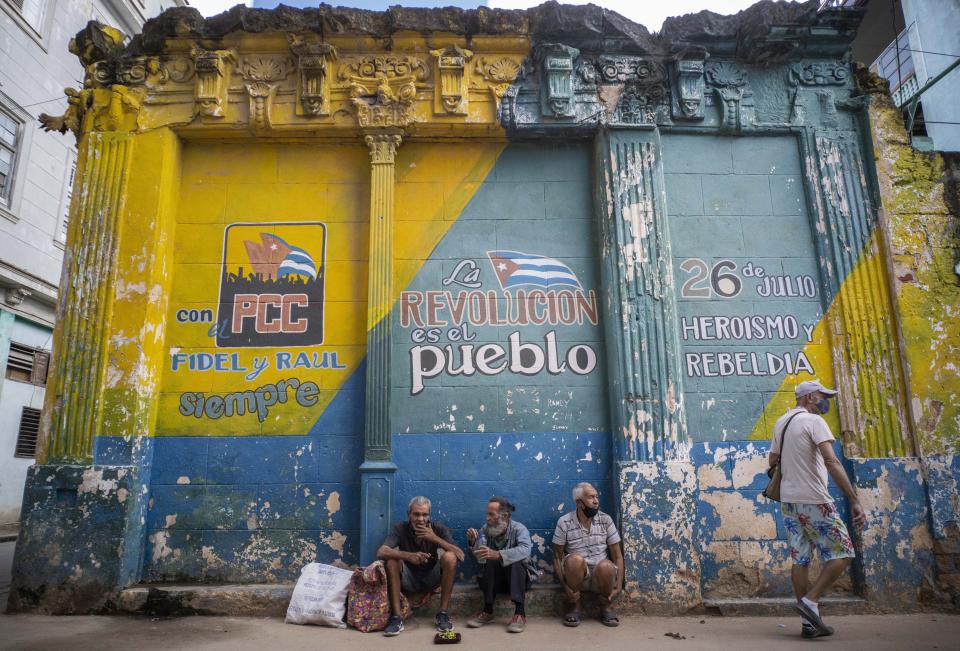 FILE - In this Dec. 23, 2020 file photo, people wearing ill-fitting masks used against the spread of the new coronavirus sit on a sidewalk next to a wall with political slogans in Havana, Cuba. In 2021, the government is implementing a deep financial reform that reduces subsidies, eliminates a dual currency that was key to the old system, and raises salaries, in hopes of boosting productivity to help alleviate an economic crisis and reconfigure a socialist system that will still grant universal benefits such as free health care and education. (AP Photo/Ramon Espinosa, File)