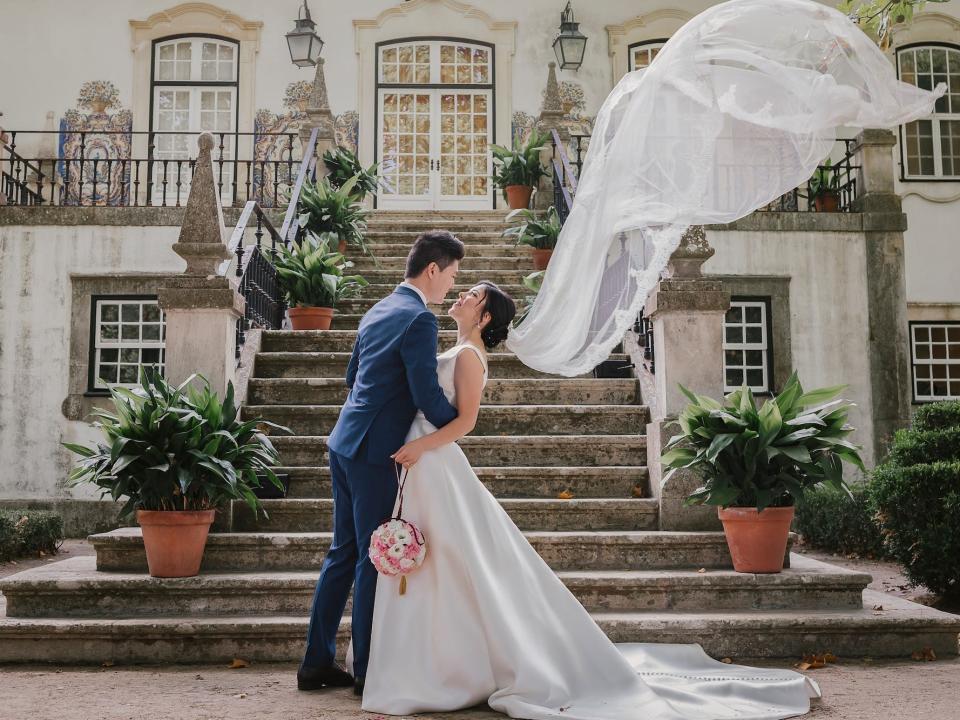 A bride and groom embrace in front of a grand staircase. The bride's veil sweeps upwards dramatically.