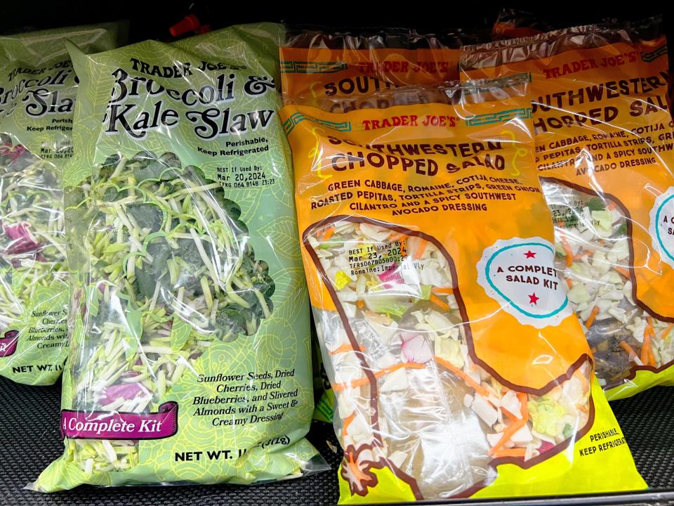 Green and orange bags of salad mix on shelves at Trader Joe's. One bag contains broccoli and kale slaw and the other has an image of a cowboy boot and contains a Southwestern mix