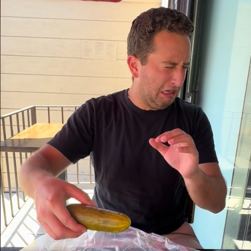 Man in a restaurant reacts humorously while holding a pickle; his expression suggests surprise or dislike