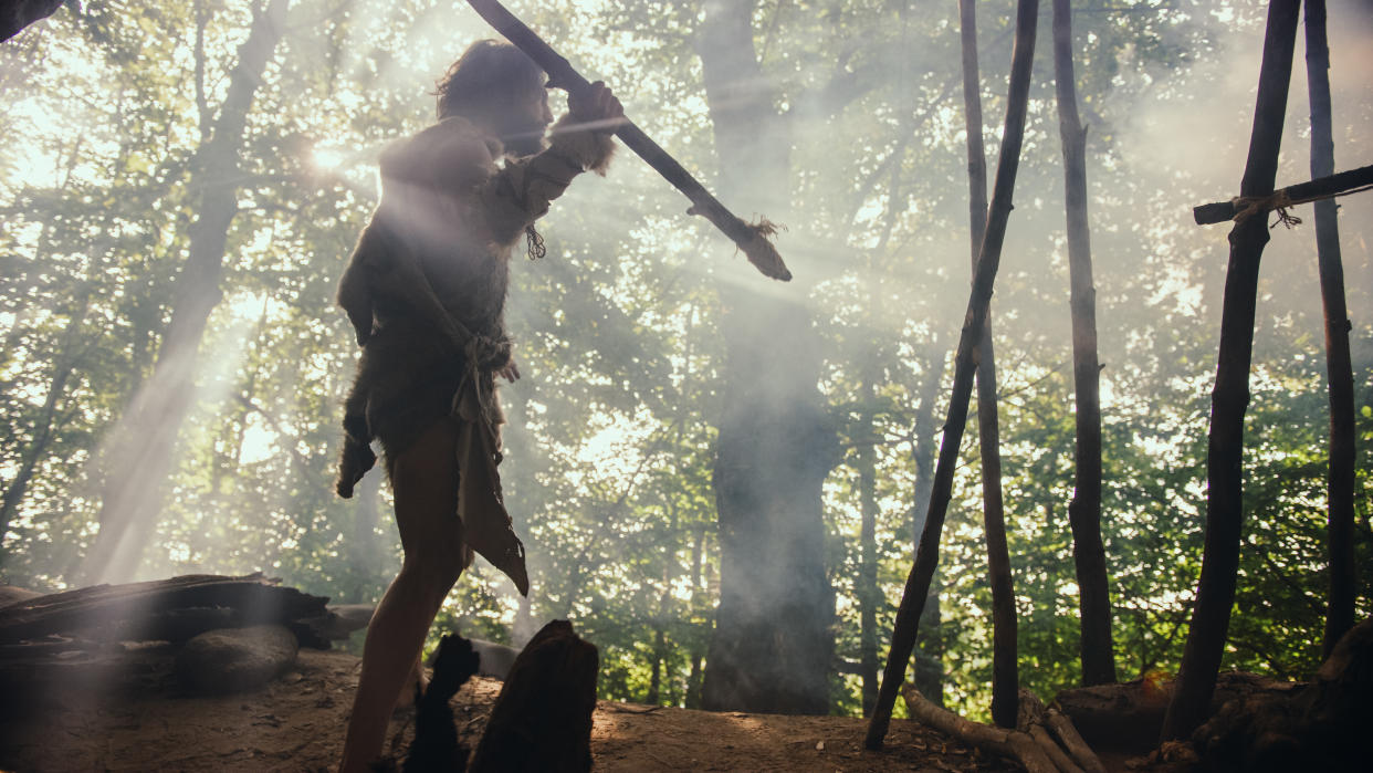 Primeval Caveman Wearing Animal Skin Holds Stone Tipped Spear, Stands at the Cave Entrance Looking over Prehistoric Forest Ready to Hunt Animal Preys. Neanderthal Going Hunting in the Jungle