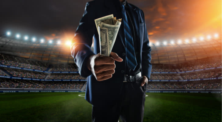 A person wearing a suit and tie holds a handful of dollar bills in the middle of a brightly lit sports stadium; representative of sports betting