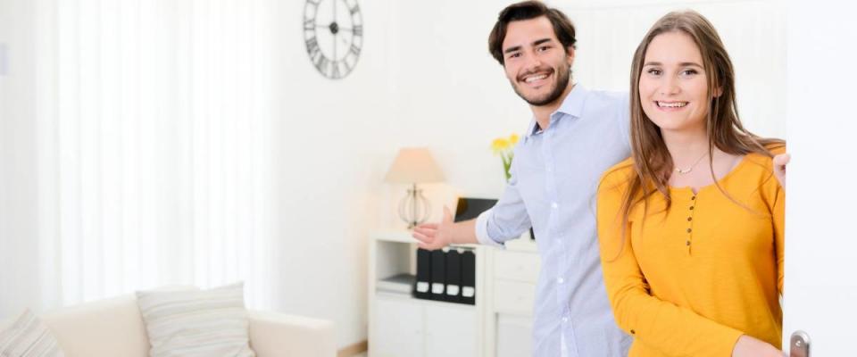 cheerful young couple man and woman welcome friends at open front door new student home apartment