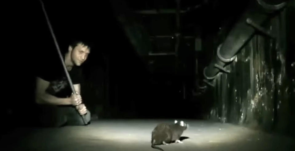 Man in a dark room holding a stick, preparing to hit a rat illuminated by a spotlight