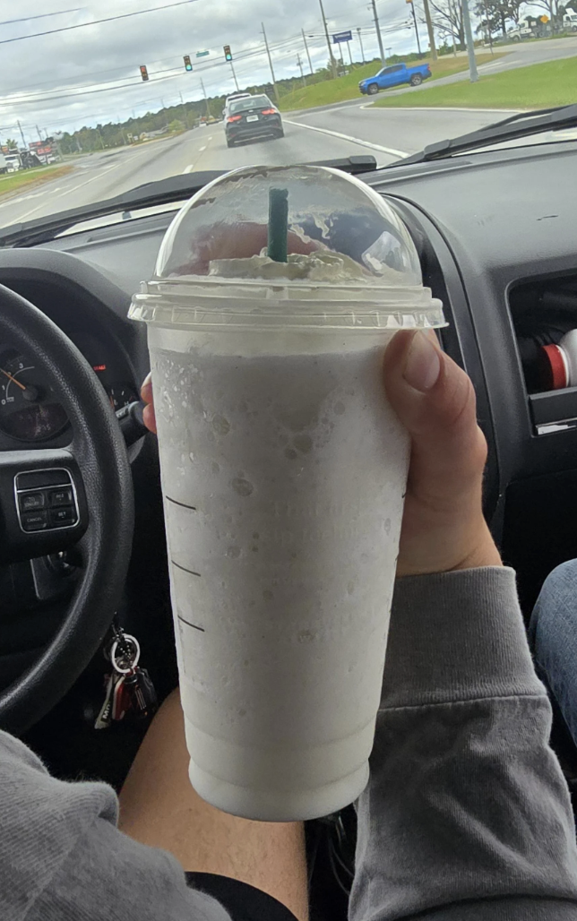 Person holding a large frosted beverage in a car, with a road and vehicles visible through the windshield