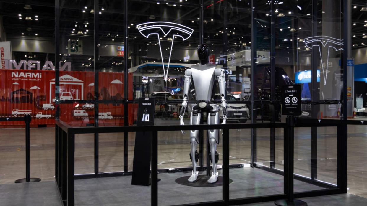  Sliver humanoid robot stands in a glass frame with a Tesla logo. 