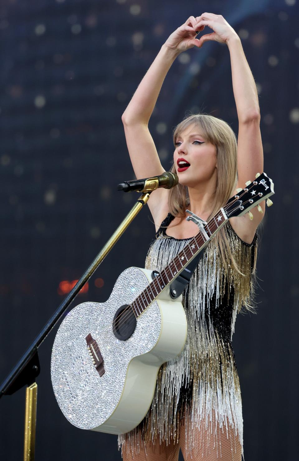 Taylor Swift on stage, wearing a fringed outfit and holding a glittery guitar, forming a heart shape with her hands above her head