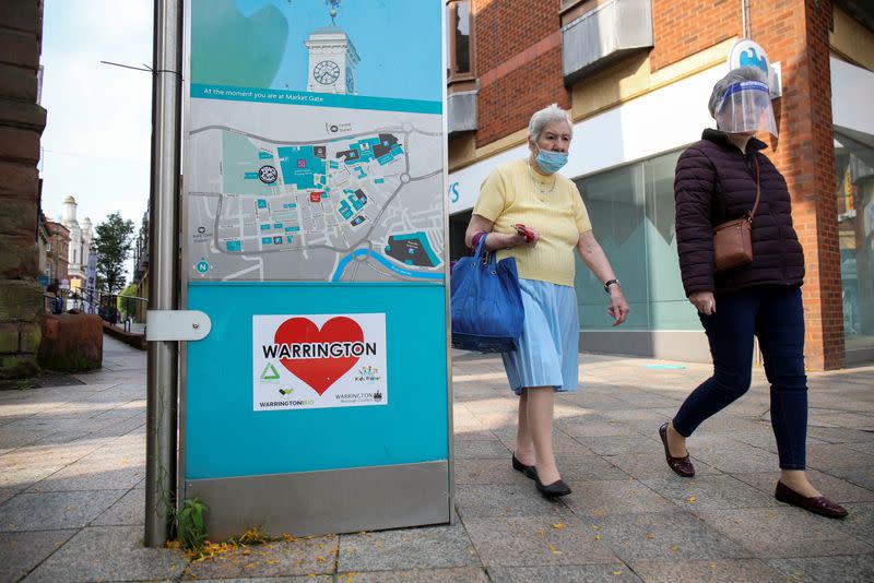 Women wearing a protective mask and a face shield walk past a city map, in Warrington