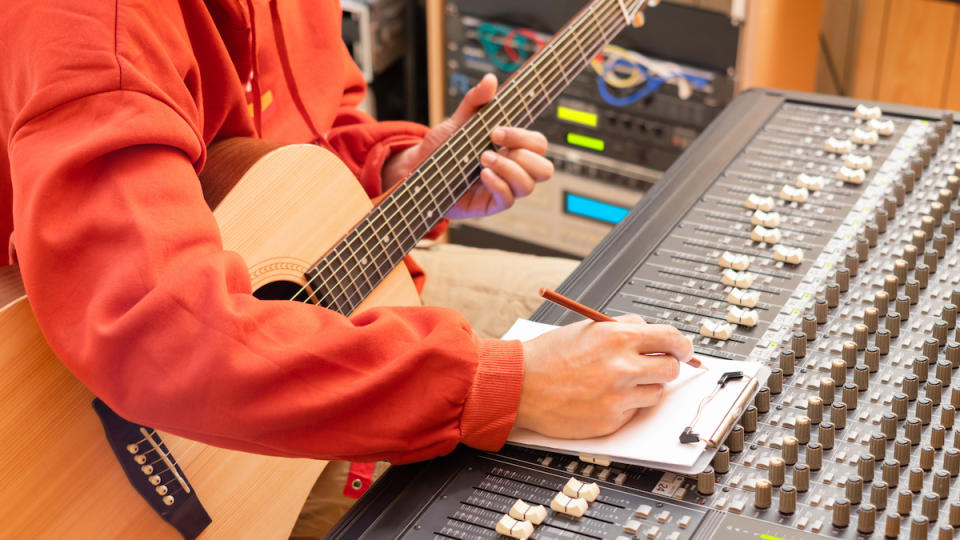 Mn in orange sweater and holding a guitar takes notes on a paper pad