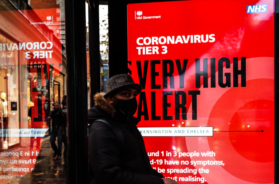 A man wearing a face mask waits beside a notice for 'Tier 3' or 'Very High' alert coronavirus restrictions displayed on the digital advertising screen of a bus stop on Kensington High Street in London, England, on December 18, 2020. Tier 3 restrictions began in London on Wednesday amid authorities' concern over escalating numbers covid-19 positive tests in the city. Shops are allowed to remain open in Tier 3 areas, but hospitality businesses are limited to takeaway and delivery service only, and entertainment venues  including theatres and cinemas must close. (Photo by David Cliff/NurPhoto via Getty Images)