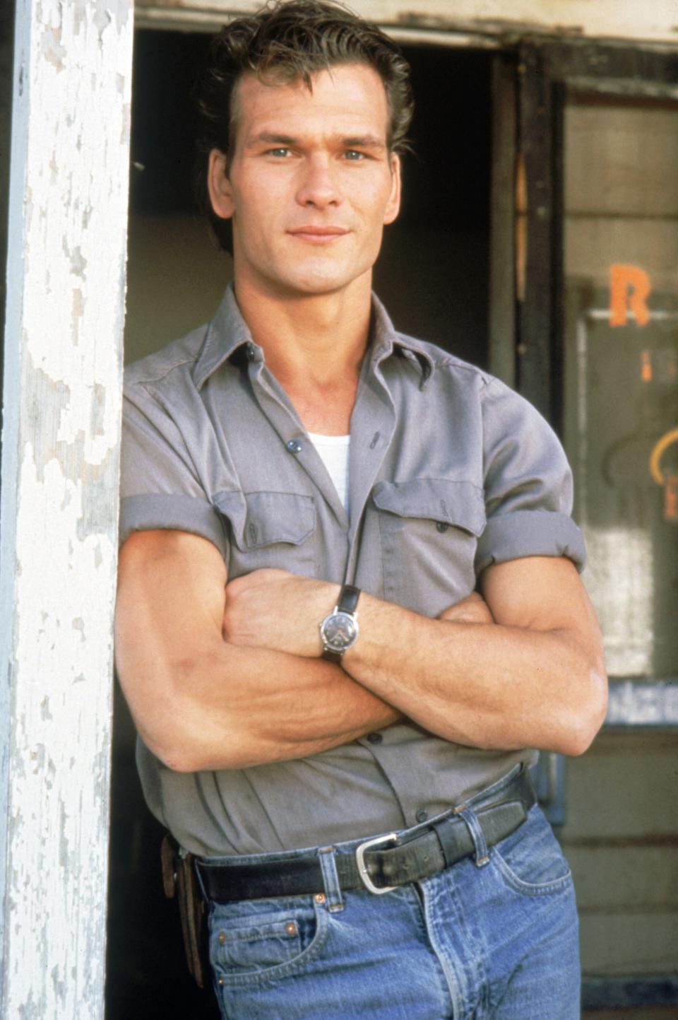 A person standing with arms crossed, wearing a short-sleeve button-up shirt and jeans, in front of a door with chipped paint