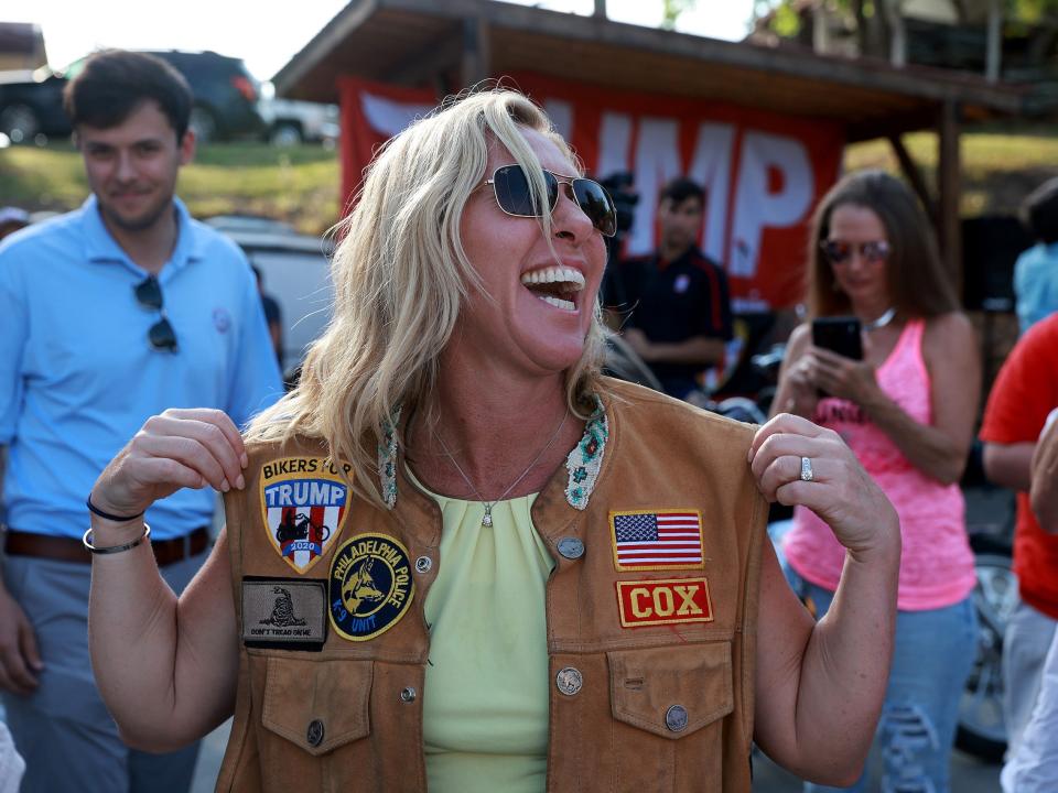Marjorie Taylor Greene wearing a biker vest in front of a big Trump sign.