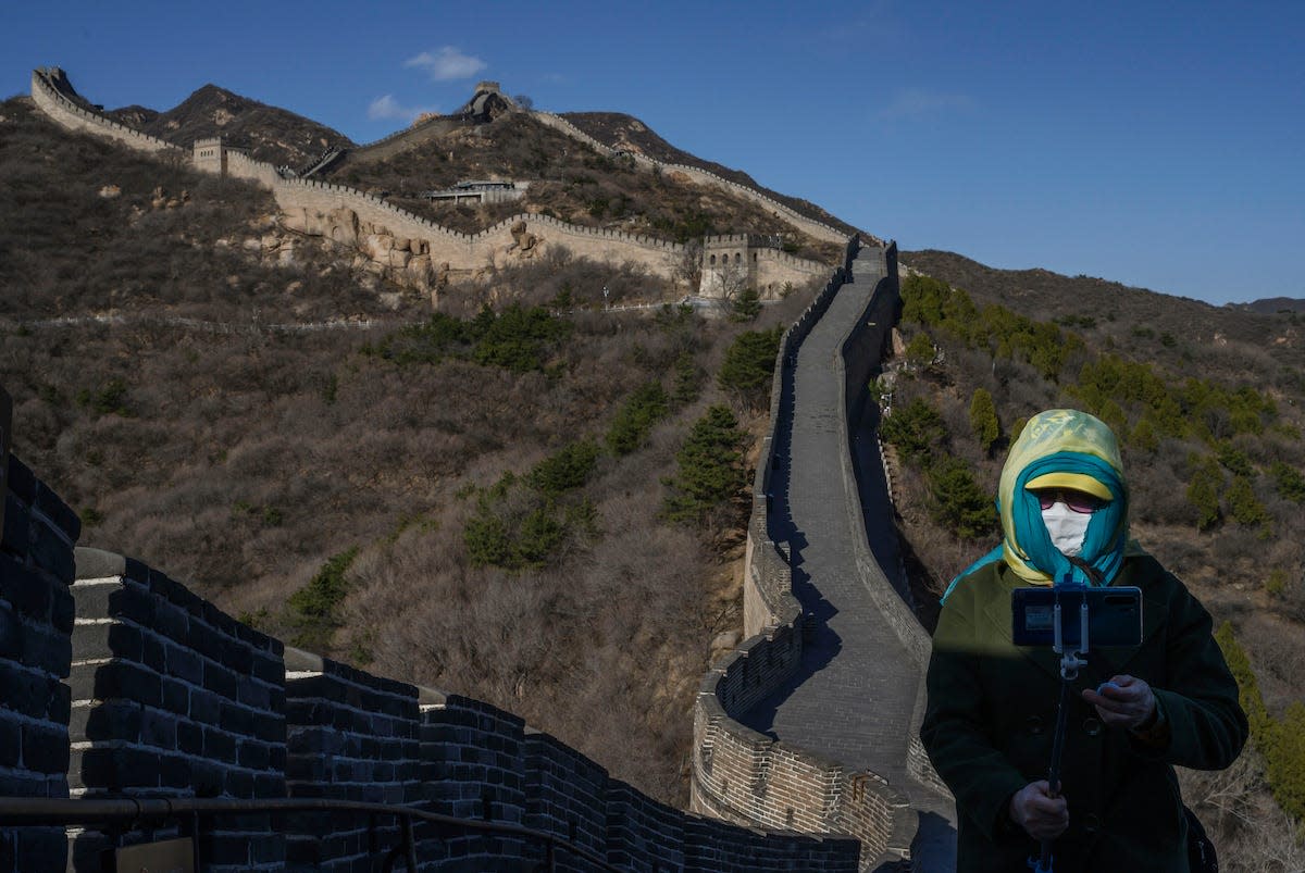 A person with a mask, headscarf and sunglasses holding a phone with a selfie stick in front of the Great Wall of China.