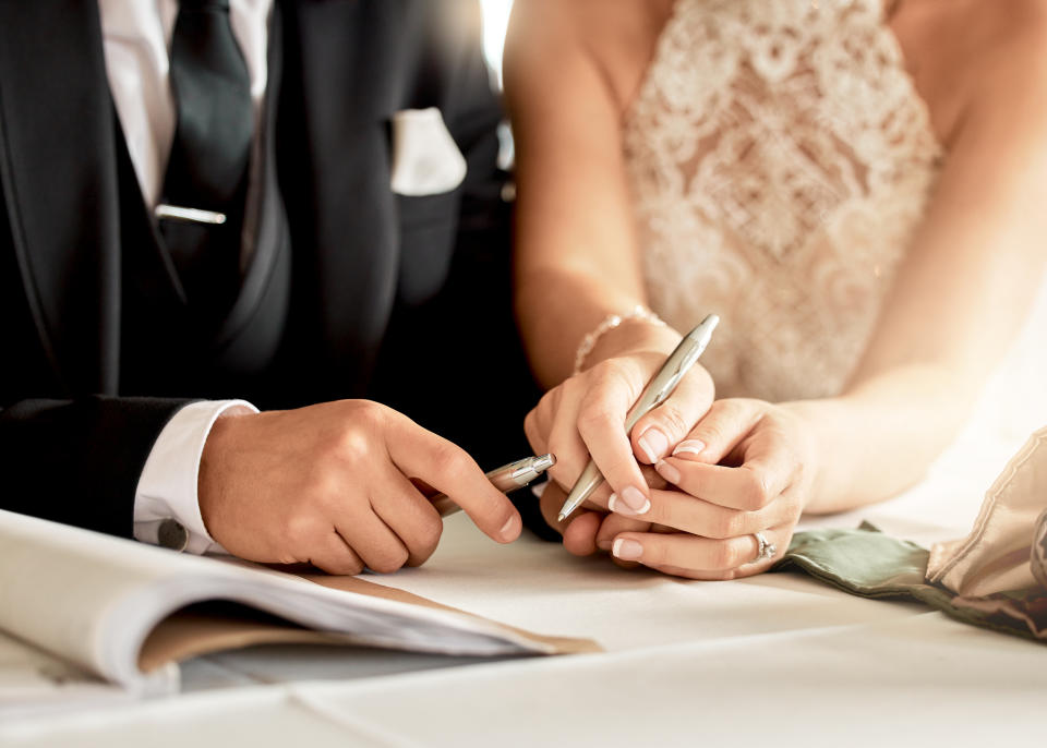 A couple in wedding attire signs a document. The groom is in a suit, and the bride is in a lace dress. Their hands and pens are the focus